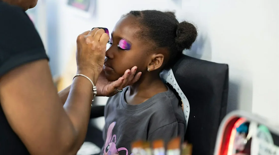 Carnivelle painting a purple design on a Black girl child sitting in a directors chair.