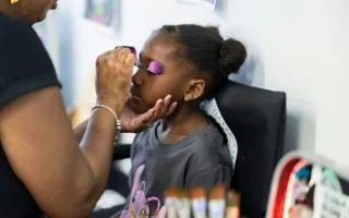 Carnivelle painting a purple design on a Black girl child sitting in a directors chair.