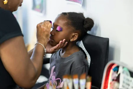Carnivelle painting a purple design on a Black girl child sitting in a directors chair.