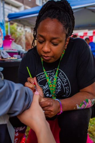 A Black face painter wearing a black uniform, with long braided hair in a pony tail, paints the arm of an out of shot child.